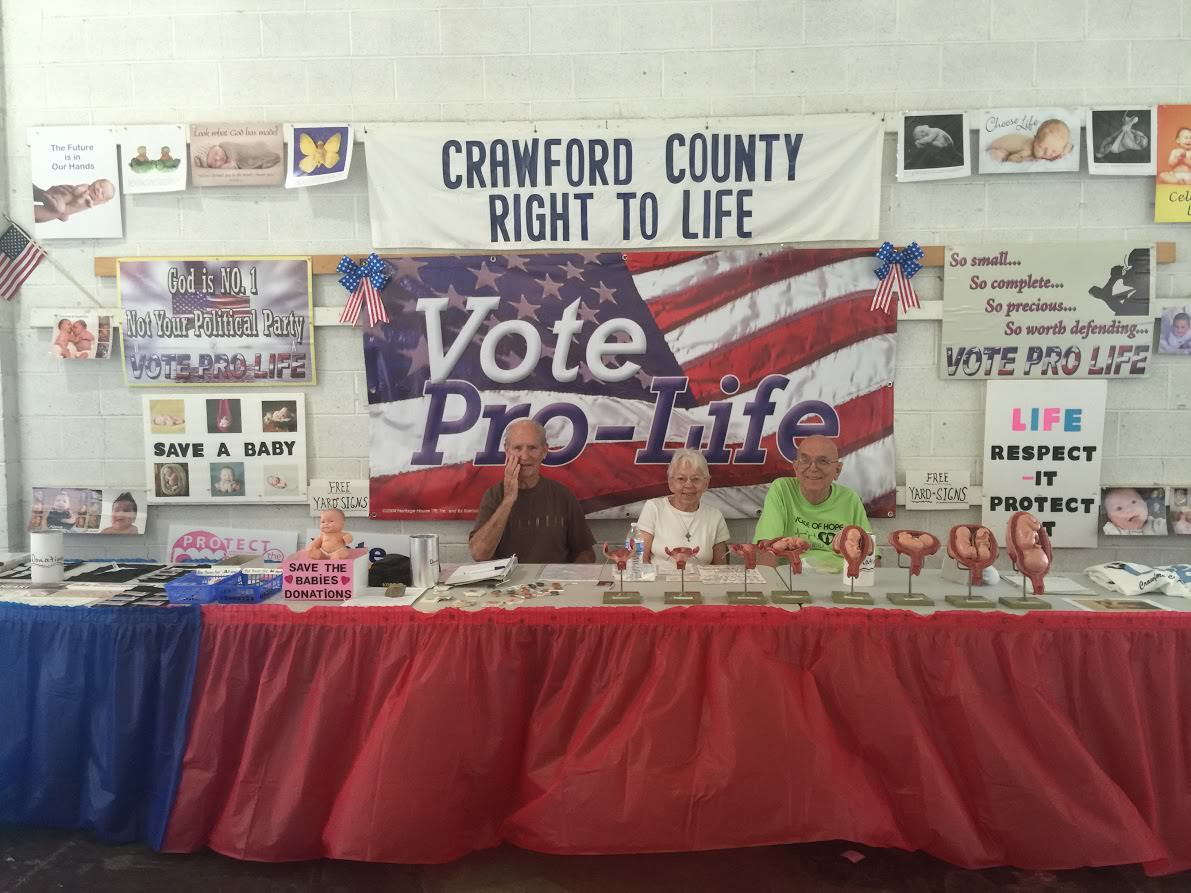 Dick and Dolly Hatfield (left) and Gary Bishop (right) sit at a booth for Crawford County Right to Life at the Crawford County Fair. (Photo: Rob Bluey/The Daily Signal)