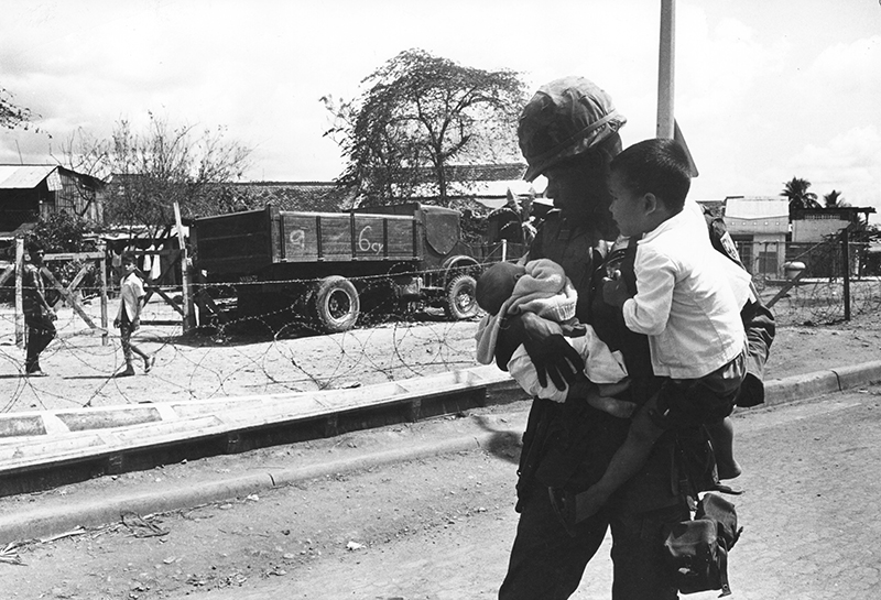 An American soldier is keeping a young boy and a newborn in his arms. In the background, a truck and other two children behind a barbed-wire fence. Saigon, 1968. (Photo: monhistorypix/Newscom)