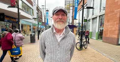 Man in sweater and ball cap with white beard stands on busy street