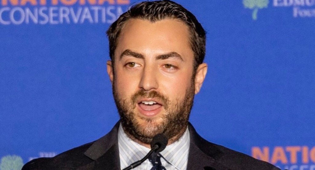 Josh Hammer in a suit in front of a blue background.