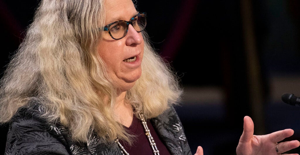 Man with long blond hair and glasses speaks in front of a microphone