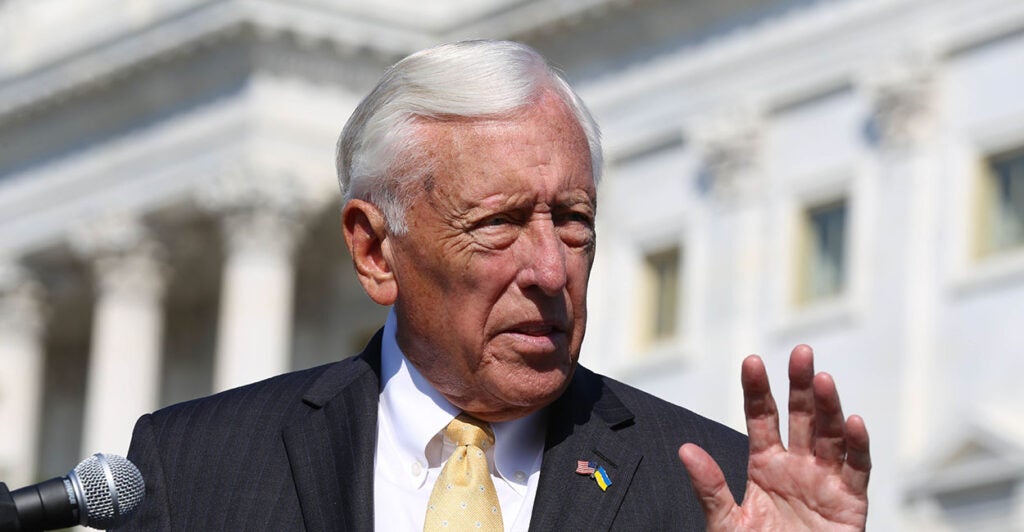 White man with silver hair in a black suit speaks in front of the Capitol building