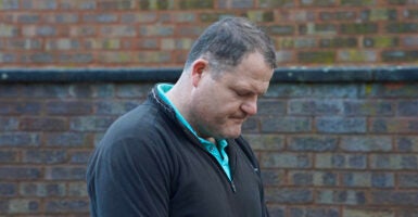 Man in a shirt and sweater bows head in front of a brick wall.