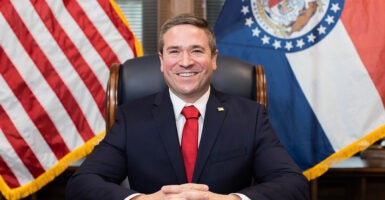 Missouri Attorney General Andrew Bailey smiles in a blue suit with a red tie in front of flags.