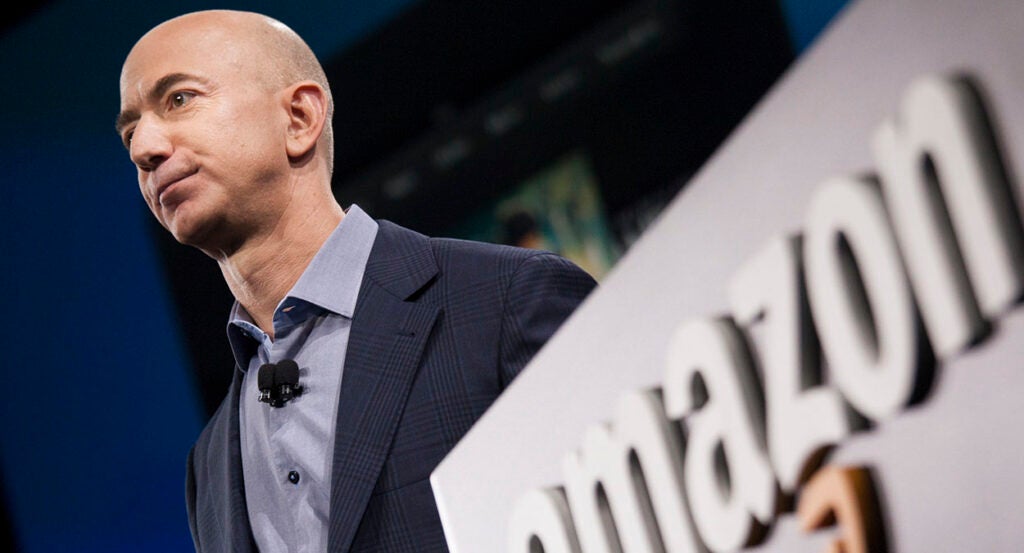 Amazon CEO Jeff Bezos in a suit near an Amazon logo.