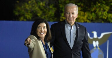 Black-haired woman in tan suit points as white-haired man in black suit stares blankly.