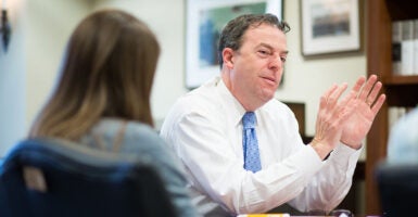 Matthew Spalding in a white shirt and a blue tie gestures in front of pictures and a bookcase