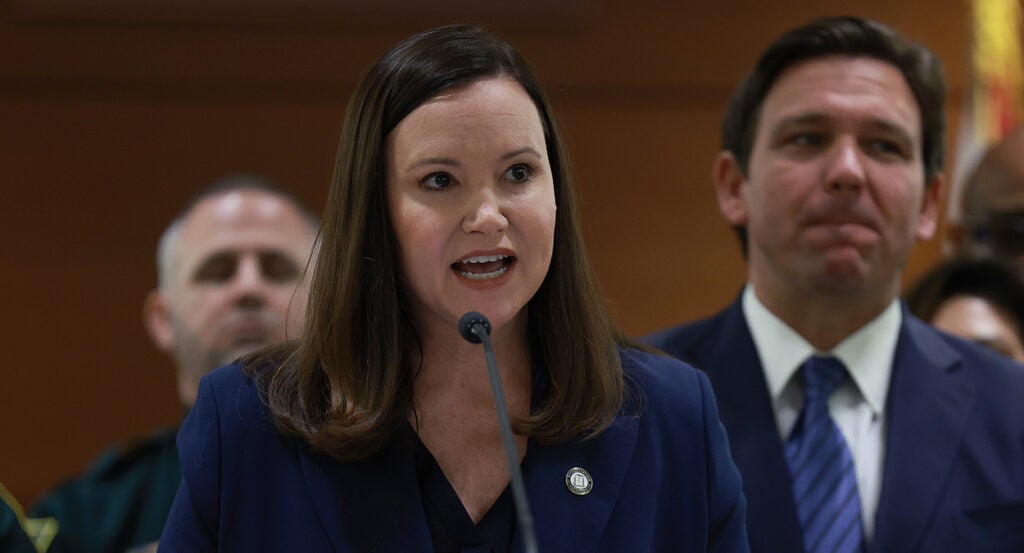 Ashley Moody in a blue suit speaks in front of Ron DeSantis in a blue suit.