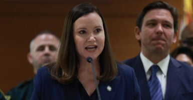 Ashley Moody in a blue suit speaks in front of Ron DeSantis in a blue suit.