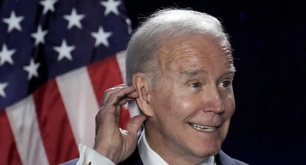 Joe Biden scratching ear in front of an American flag