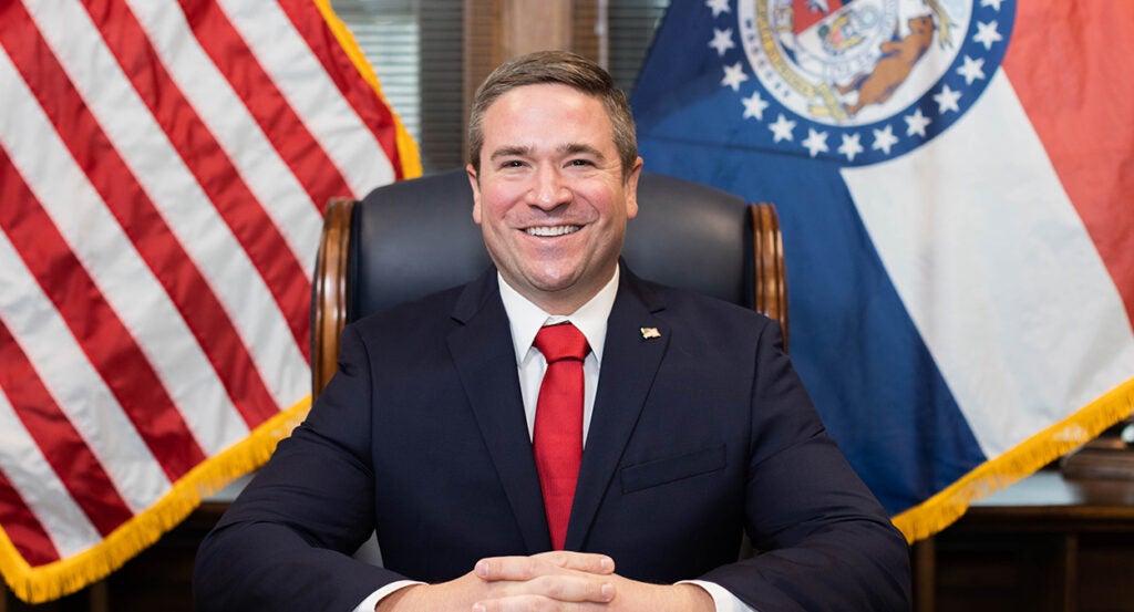 Andrew Bailey in a suit with a red tie in front of an American flag and a Missouri flag