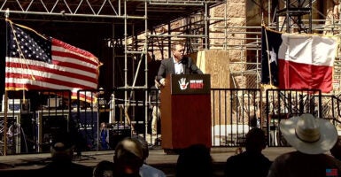 Mark Meckler speaks on a stage outside the Texas Capitol building with an American flag and a Texas flag behind him.