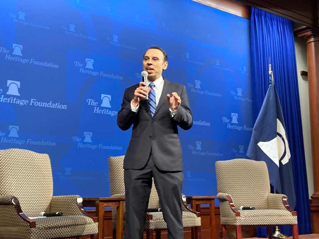 Roger Severino stands with a microphone in a suit in front of a wall with The Heritage Foundation logo