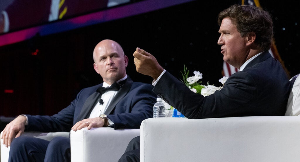 Kevin Roberts in black tie listens as Fox News host Tucker Carlson gestures.