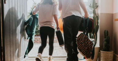 A boy and a girl are seen from behind leaving their house with backpacks and lunch boxes.