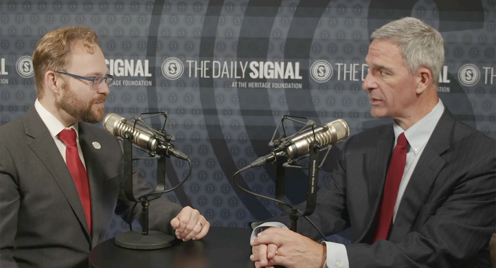 The Daily Signal's Tyler O'Neil in a grey suit and red tie listens as former Virginia Attorney General Ken Cuccinelli speaks in a pin-stripe suit and red tie