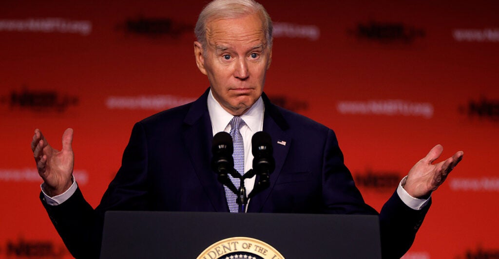 President Joe Biden speaks from stage in a suit with his hands raised and palms up.