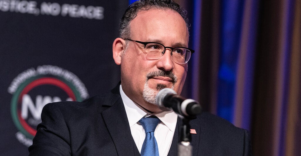 Secretary of Education Miguel Cardona speaks into a microphone wearing a suit.