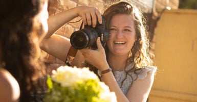 Emilee Carpenter smiles while pointing a camera at a bride holding flowers