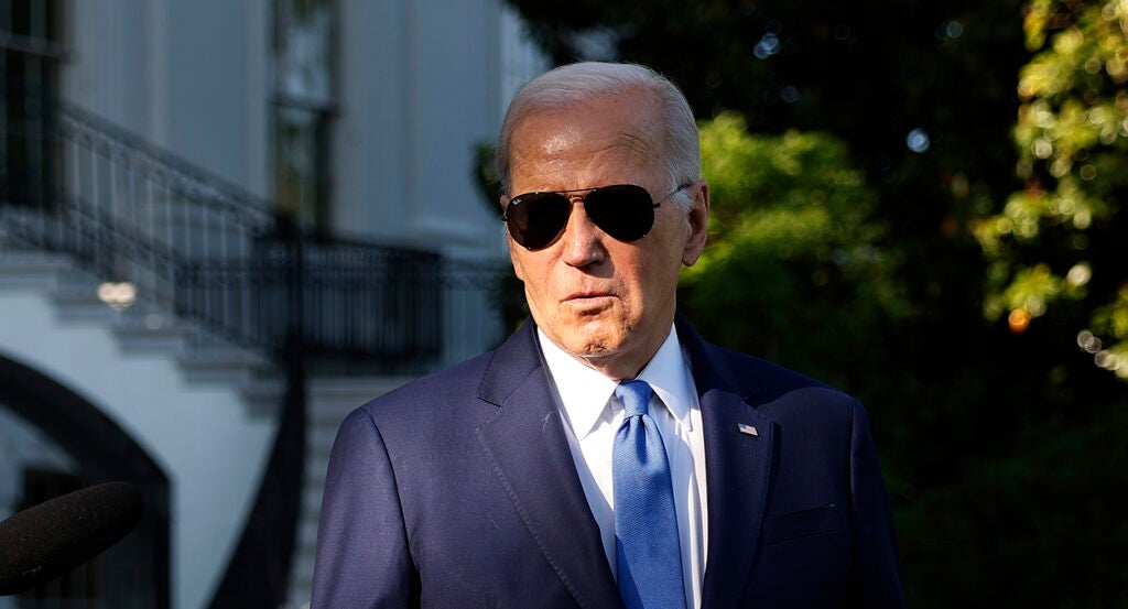 Joe Biden stands with dark sunglasses in a blue suit in front of the White House