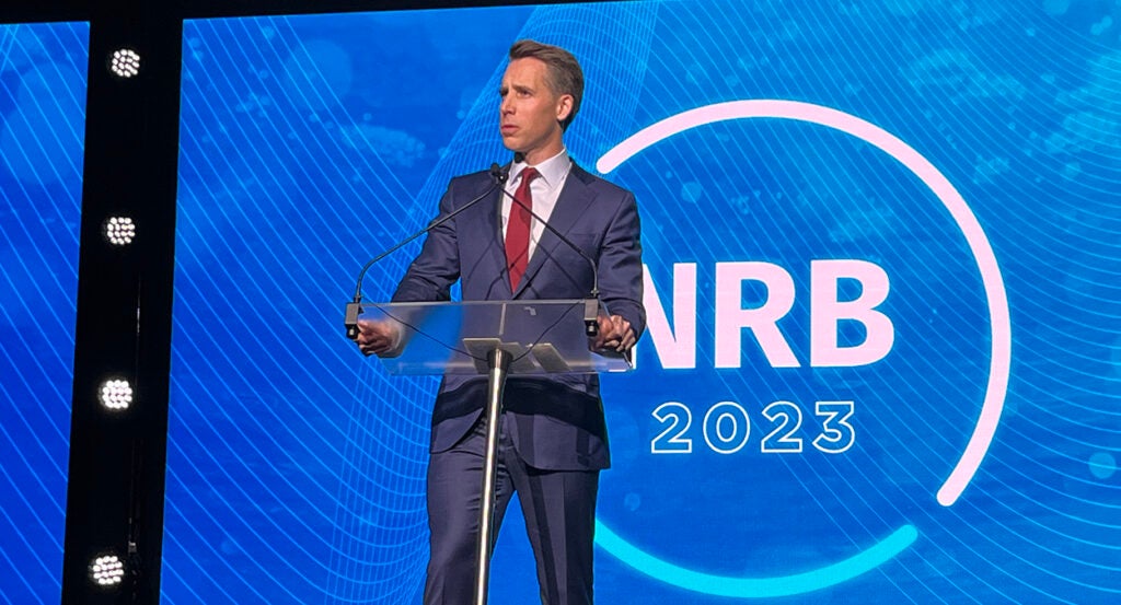 Senator Josh Hawley in a blue suit with a red tie speaks in front of the NRB 2023 logo