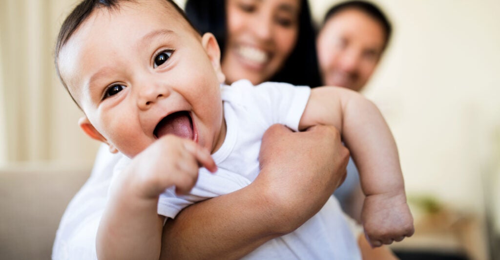 baby boy smiles at camera with parents in background