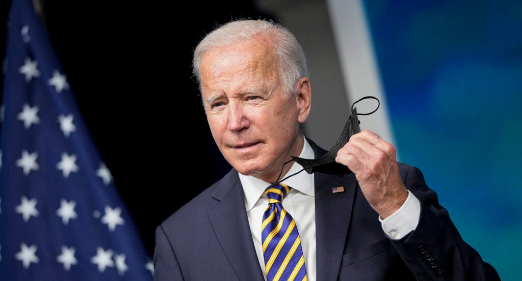 Joe Biden in a suit with an American flag pin holds a black mask in his left hand.