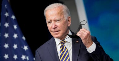 Joe Biden in a suit with an American flag pin holds a black mask in his left hand.