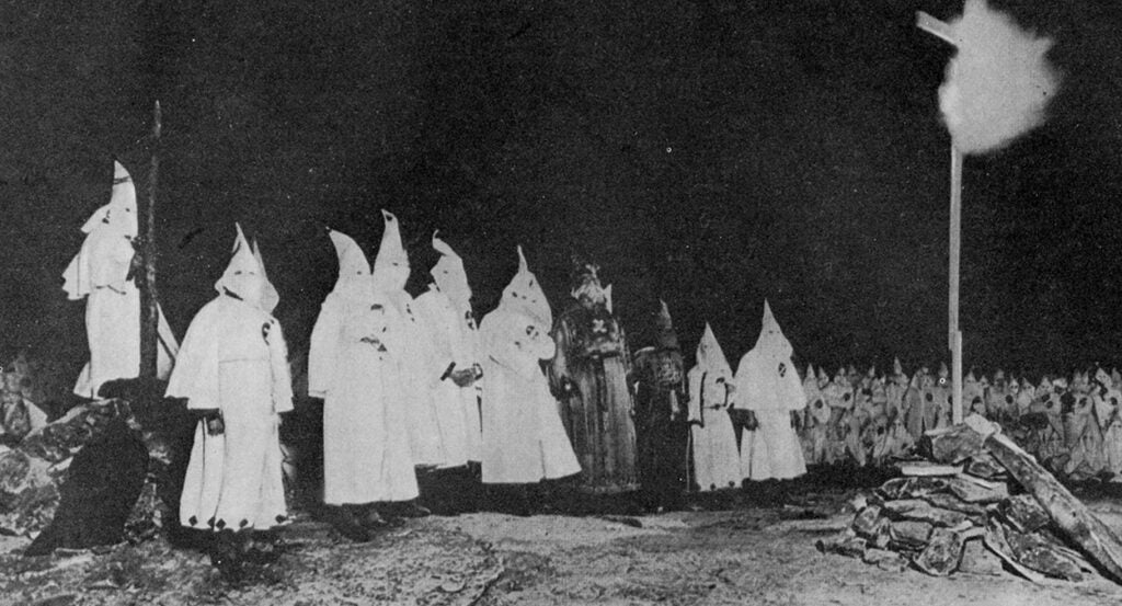 Men in white KKK hoods stand behind a burning cross.