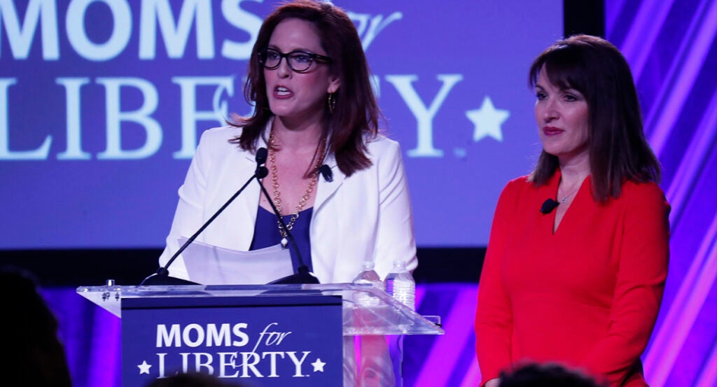 Tiffany Justice in a blue dress and white blazer speaks with Tina Descovich in a red dress in front of a Moms for Liberty logo