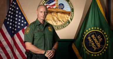 Jason Owens stands in his uniform in front of American and Border Patrol flags.