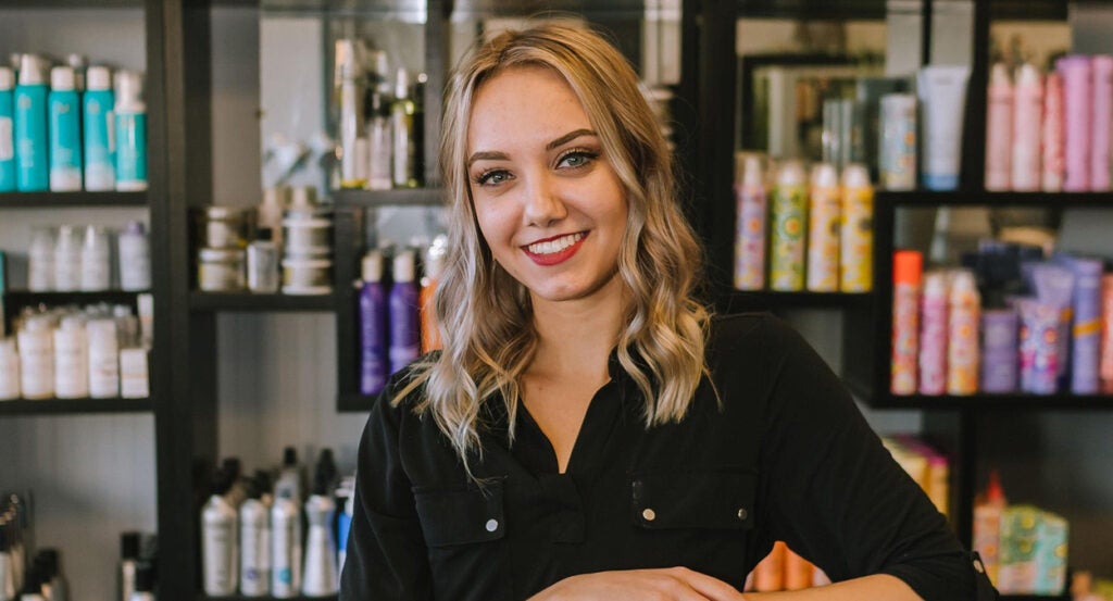 Young blonde hair stylist smiles in front of hair products