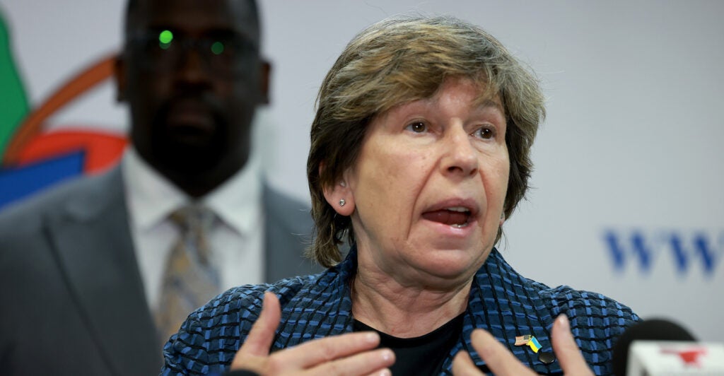 Randi Weingarten, president of the American Federation of Teachers. is speaking and using her hands at a podium. There is a man in a suit and tie standing behind her.