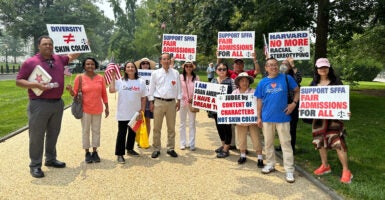 A group of Asian American men and women hold up signs in support of the Supreme Court's decision to get rid of affirmative action. They stand on a tan gravel path, in front of rows of trees.