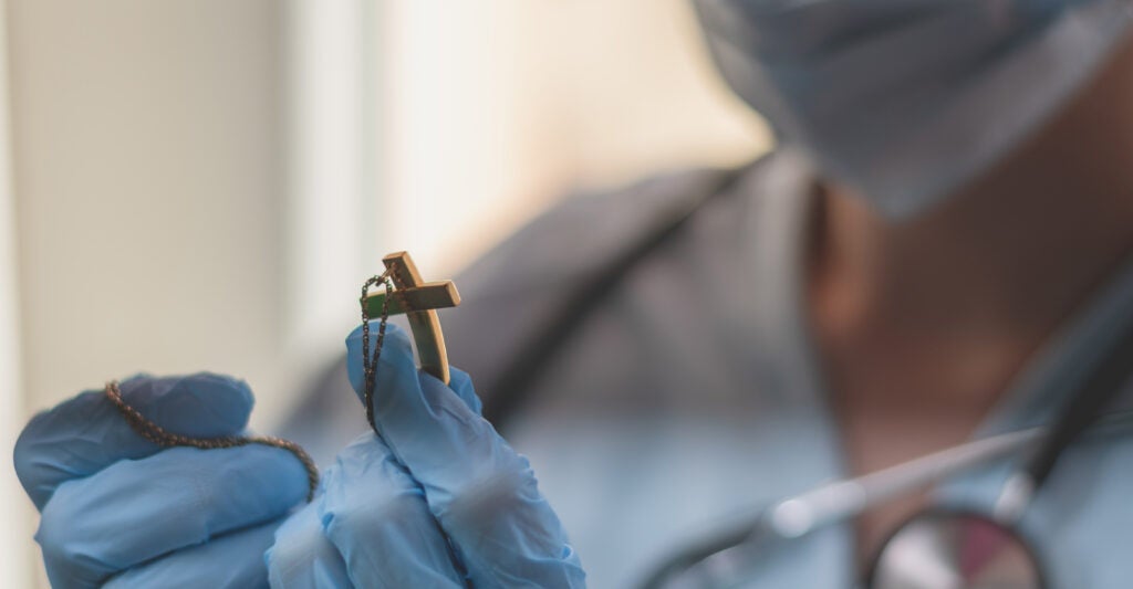 A medical professional holds the cross part of a rosary. She is wearing a mask, scrubs, and gloves. The image is a close up on her hands.