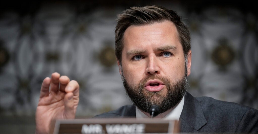 JD Vance is talking while using his hand to make a gesture. He is seated in front of a name card.