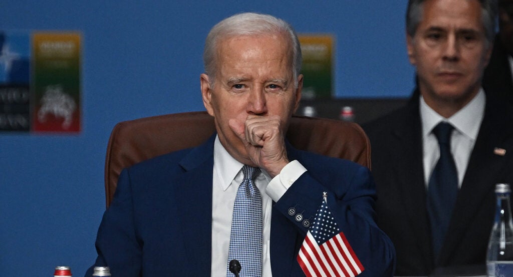 Joe Biden in a blue suit coughs with Antony Blinken looking over his shoulder