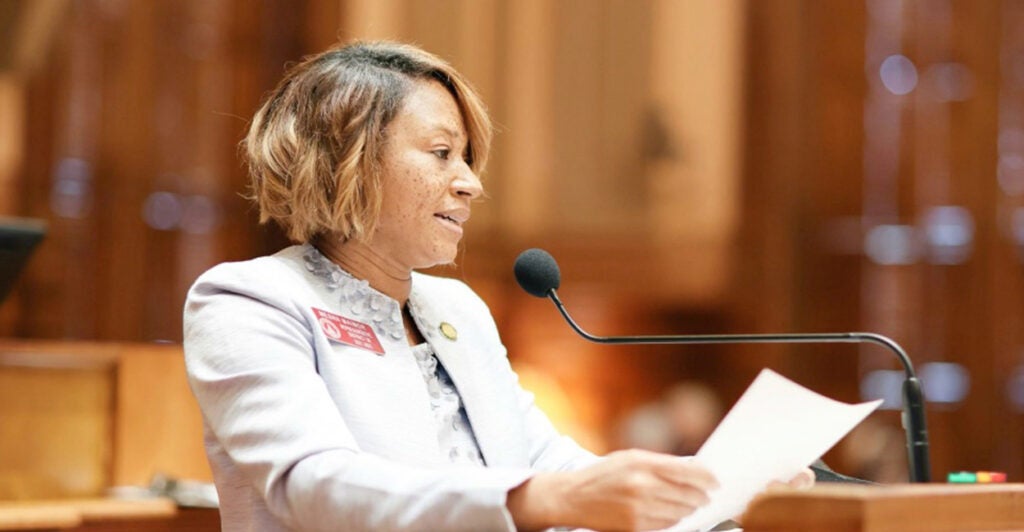 Mesha Mainor speaks into a microphone holding a paper and wearing a light suit jacket.