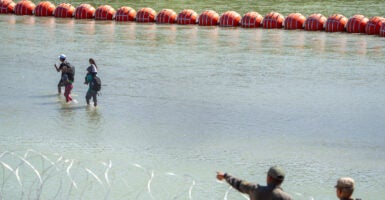 A string or red buoys are at floating on the river, with three people wading in the water and two people in the bottom right pointing at them.