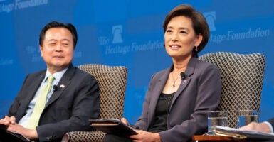 An asian man and woman sit side by side in business clothes and talk. They sit in front of a blue background.