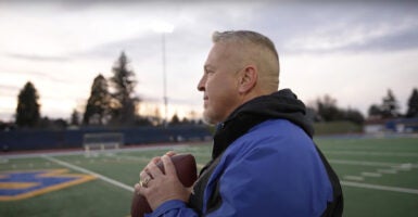 Coach Joe Kennedy stand on a football field holding a football.