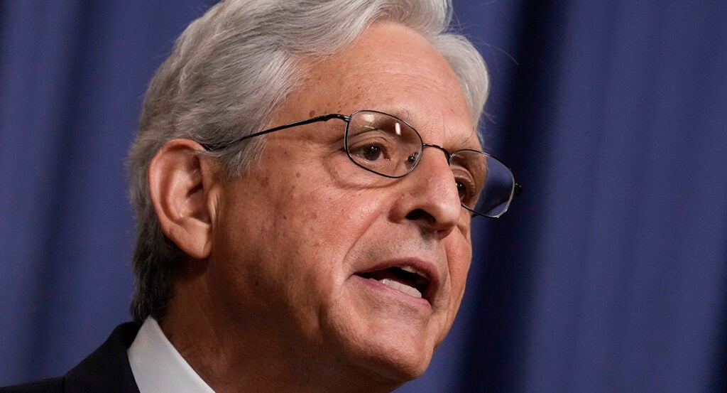 Merrick Garland with white hair in a suit and wearing glasses stands in front of a blue background