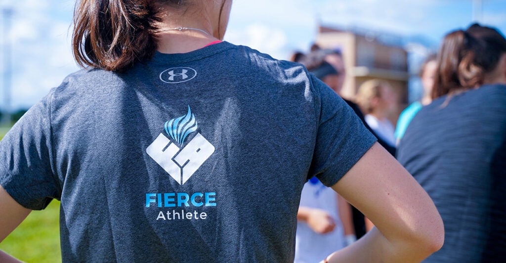 A woman stands on the edge of a sports field in a FIERCE Athlete t-shirt.