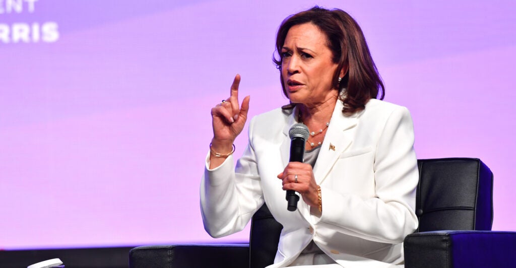 Kamala Harris wears a white blazer, holds a microphone, and speaks in front of a purple background. She is seated on a stage.