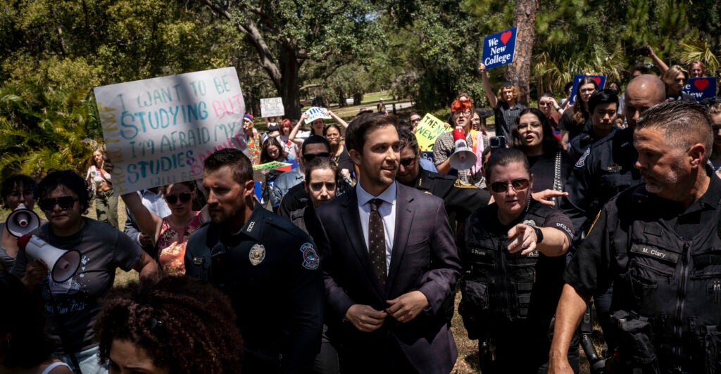 Chris Rufo makes hs way through a crowd of protestors outdoors. He is surrounded by security.