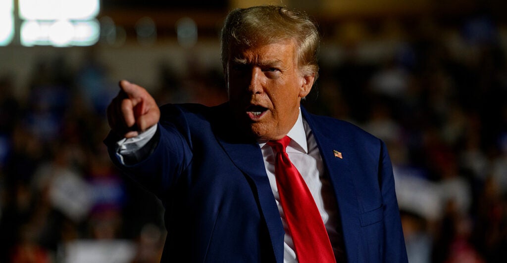 Former President Donald Trump points to the crowd in a dark blue suit and red tie during a political rally.