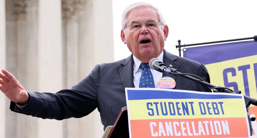 Robert Menendez in a pin-stripe suit speaks in front of a sign reading, "Student Debt Cancellation Is Legal"