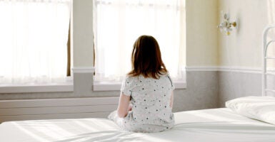 A girl sits on a bed that is covered in a white sheet and looks out the window.