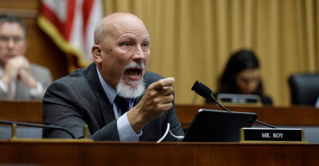 Chip Roy gestures in a suit in front of an American flag