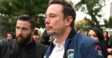 Elon Musk walks outside after leaving a meeting wearing a white shirt and dark blue jacket.